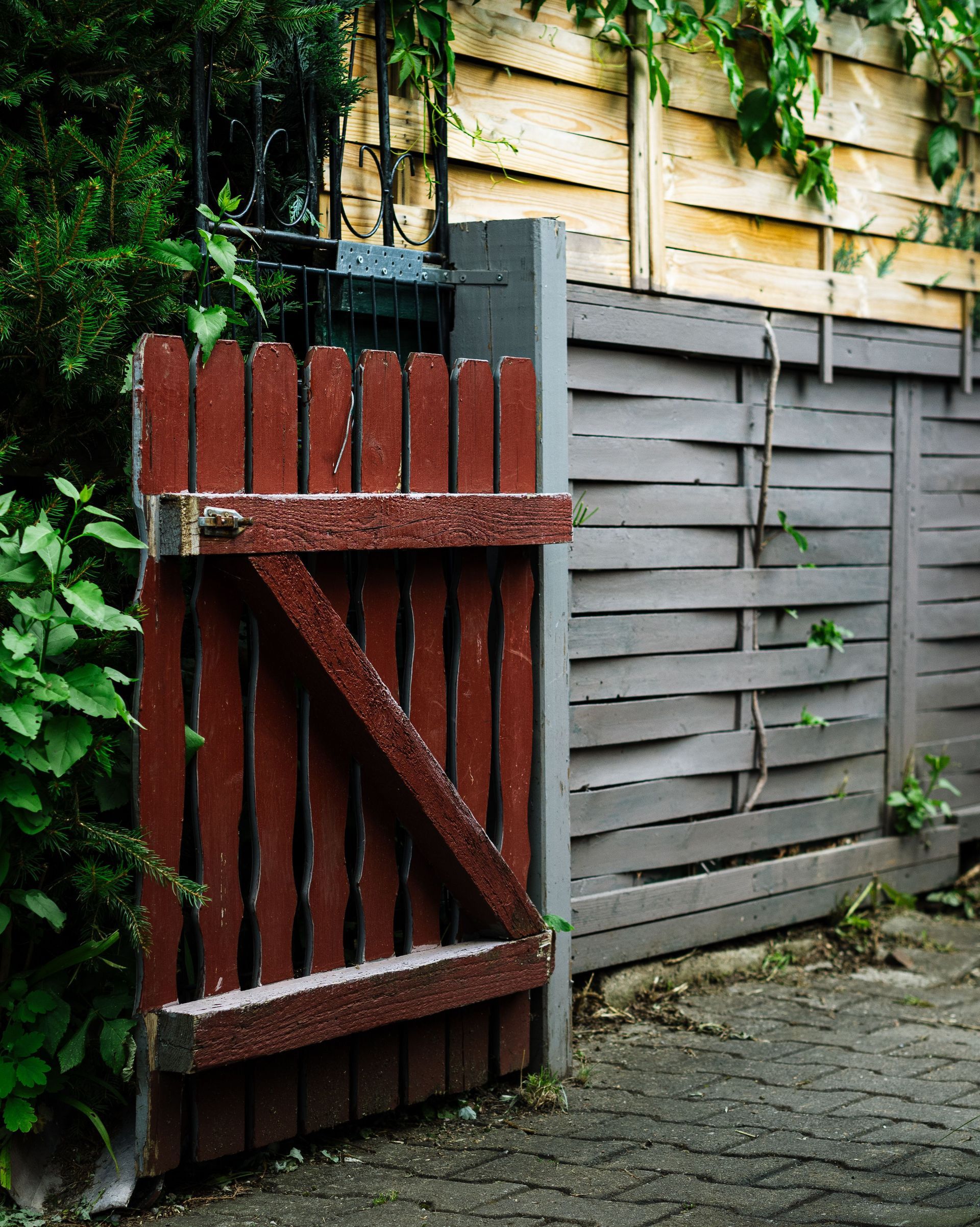 Red wooden gate with diagonal brace beside a gray fence and leafy greenery along a stone path