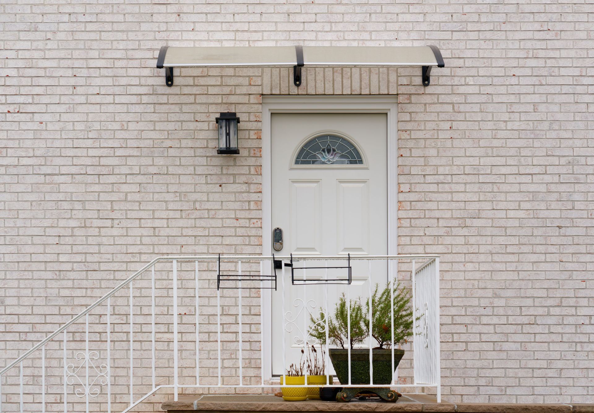 White front door with awning and black lantern on a pale brick house, behind a small white railing and potted plants