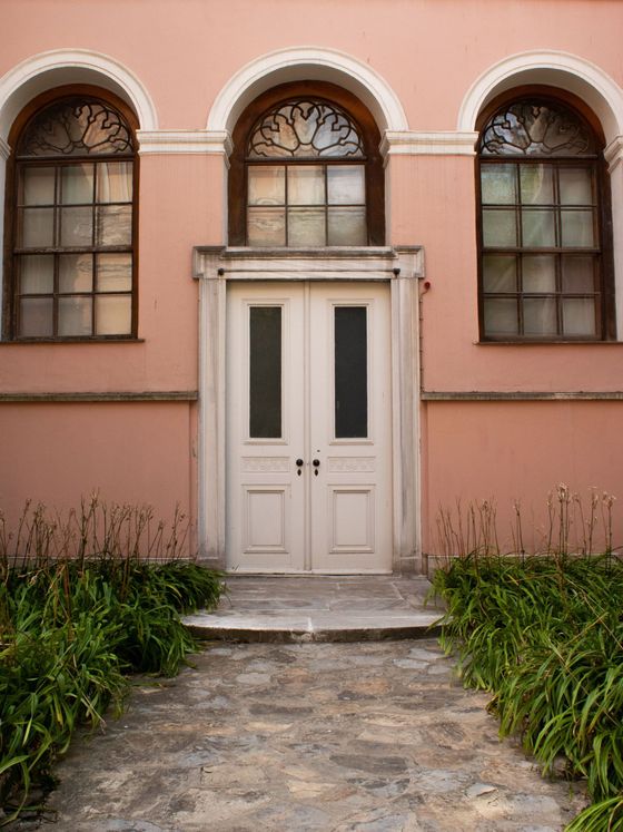 Pink building facade with arched windows and a white double door, framed by a stone path and green plants