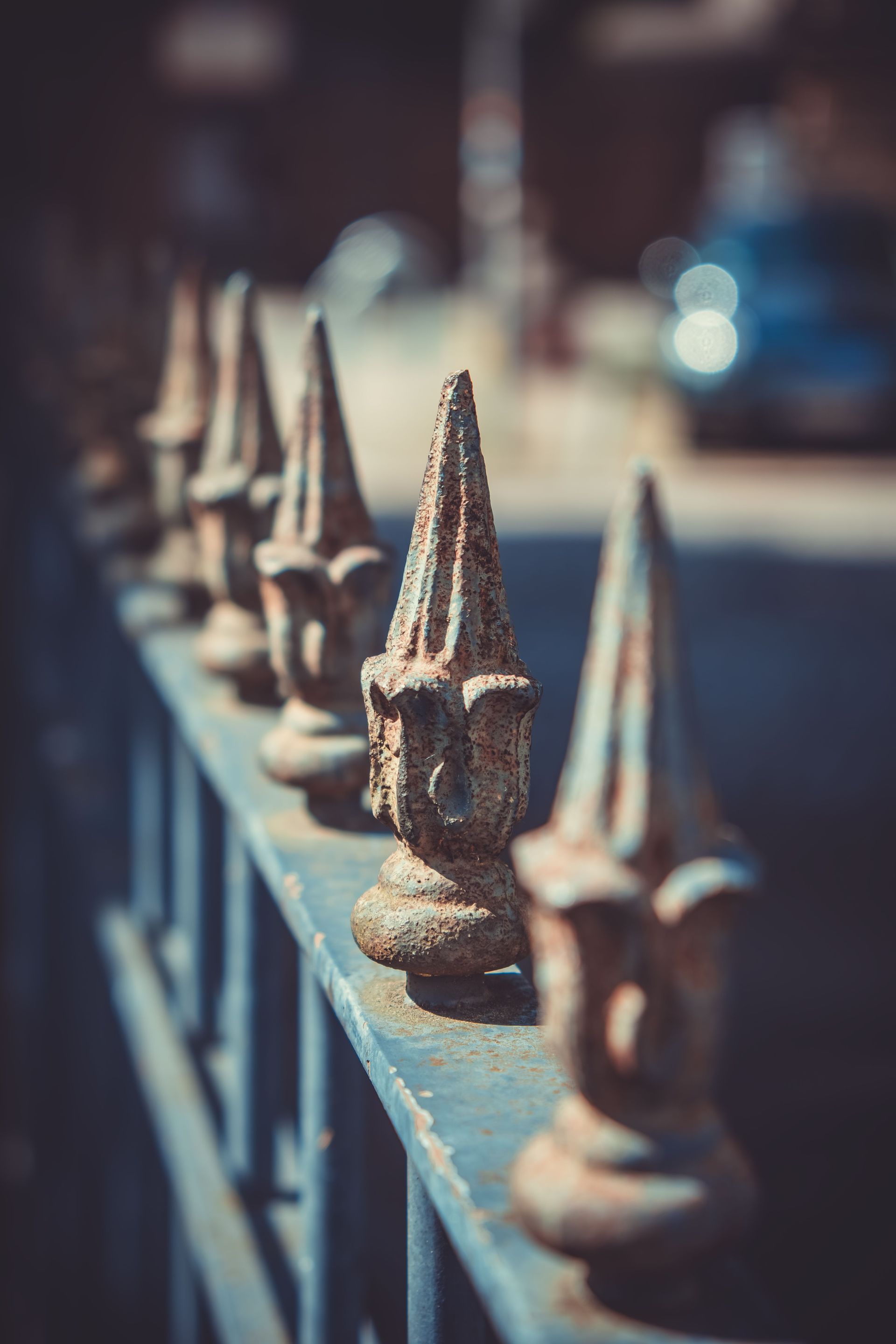 Row of weathered metal fence finials along a blurred sidewalk, with shallow depth of field