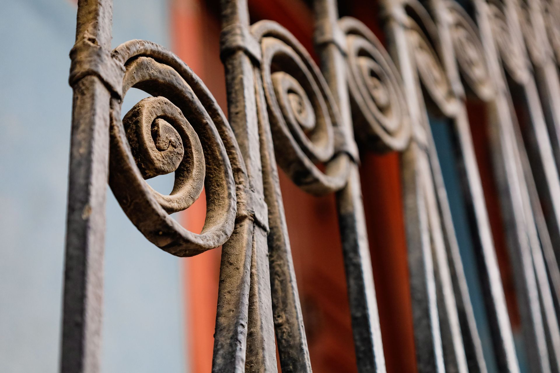 Rusty wrought-iron fence with decorative scrollwork against a blurred orange wall