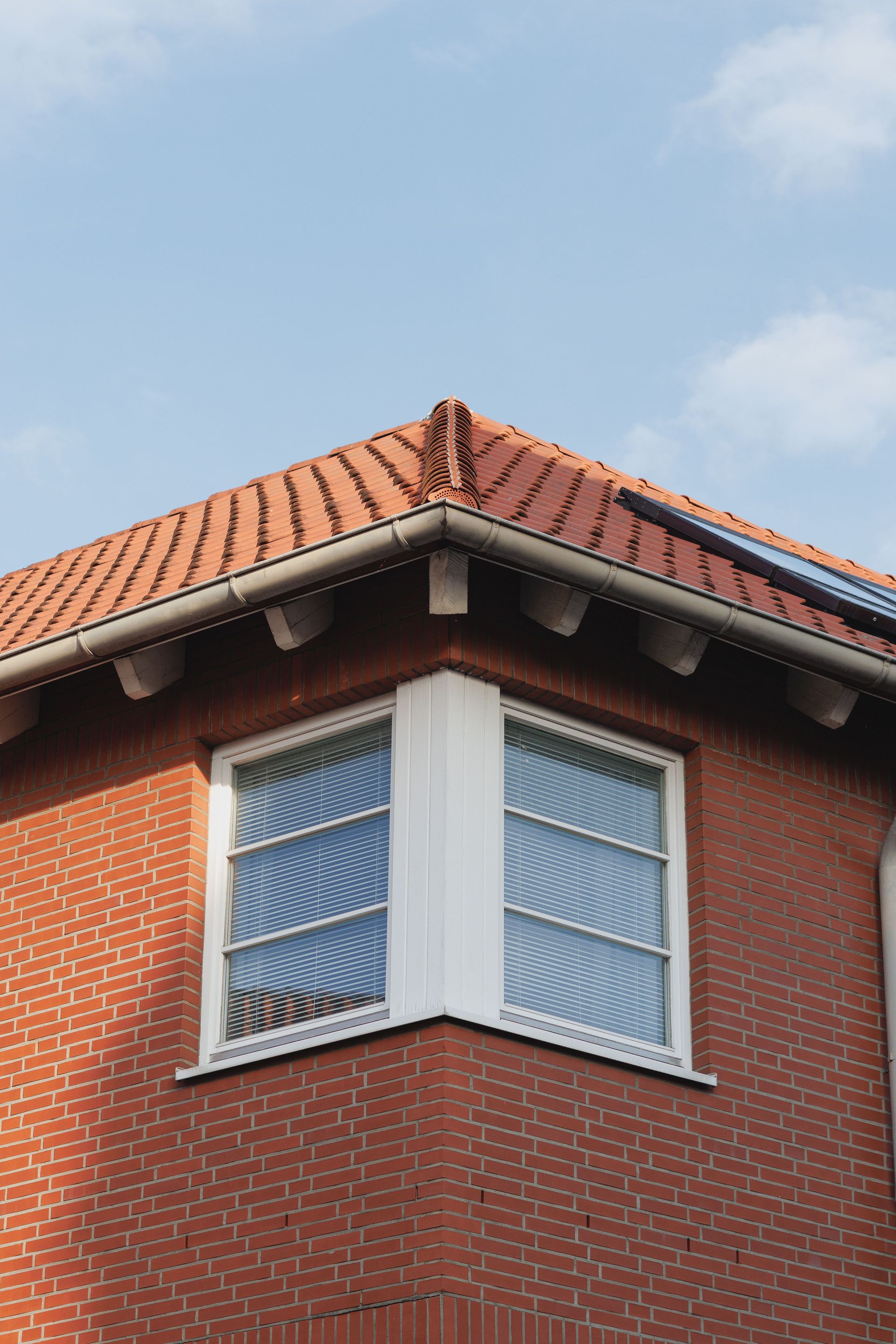 Corner of red brick building with a tiled roof and white-framed bay window against a blue sky