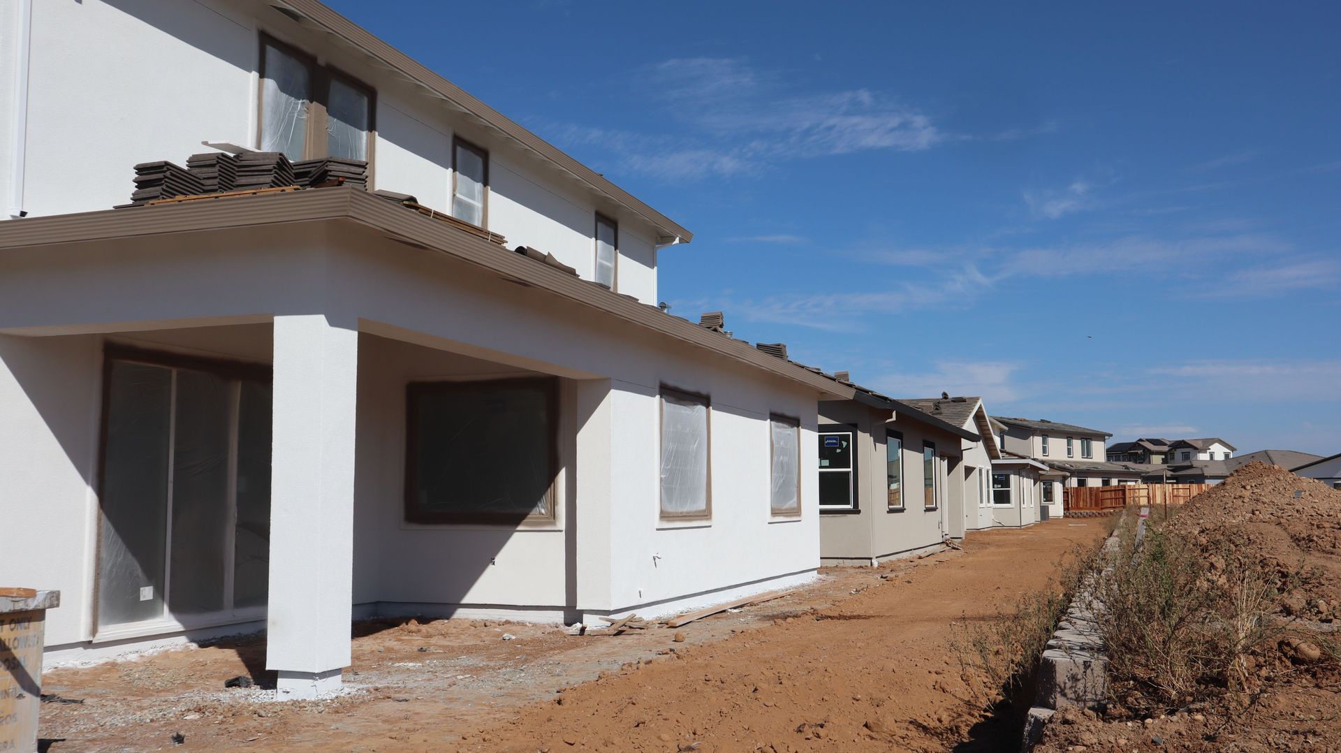 Row of unfinished white townhouses under construction beside a dirt road under a blue sky