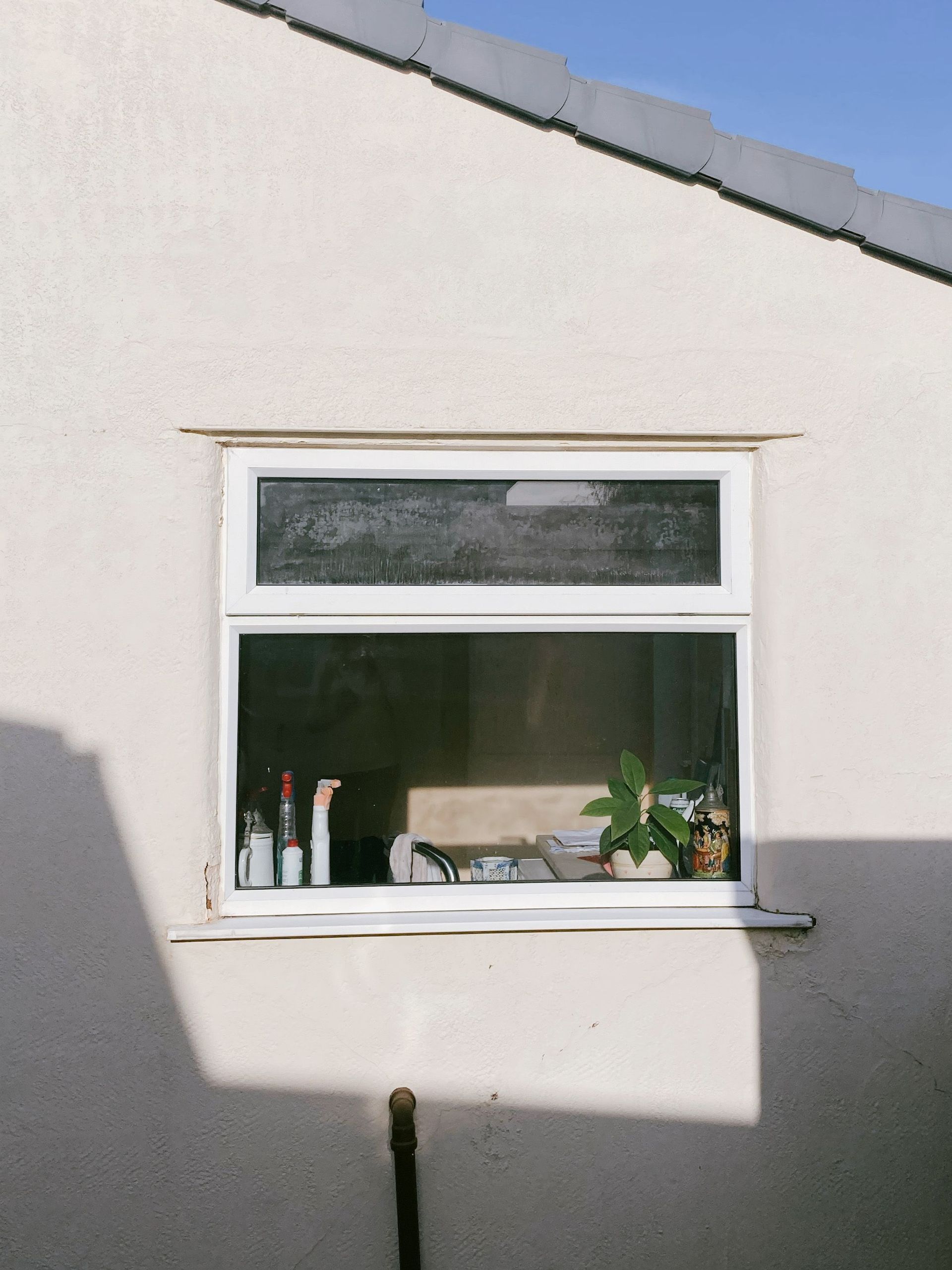 White house wall with a small window and potted plants on the sill, partly in shadow.