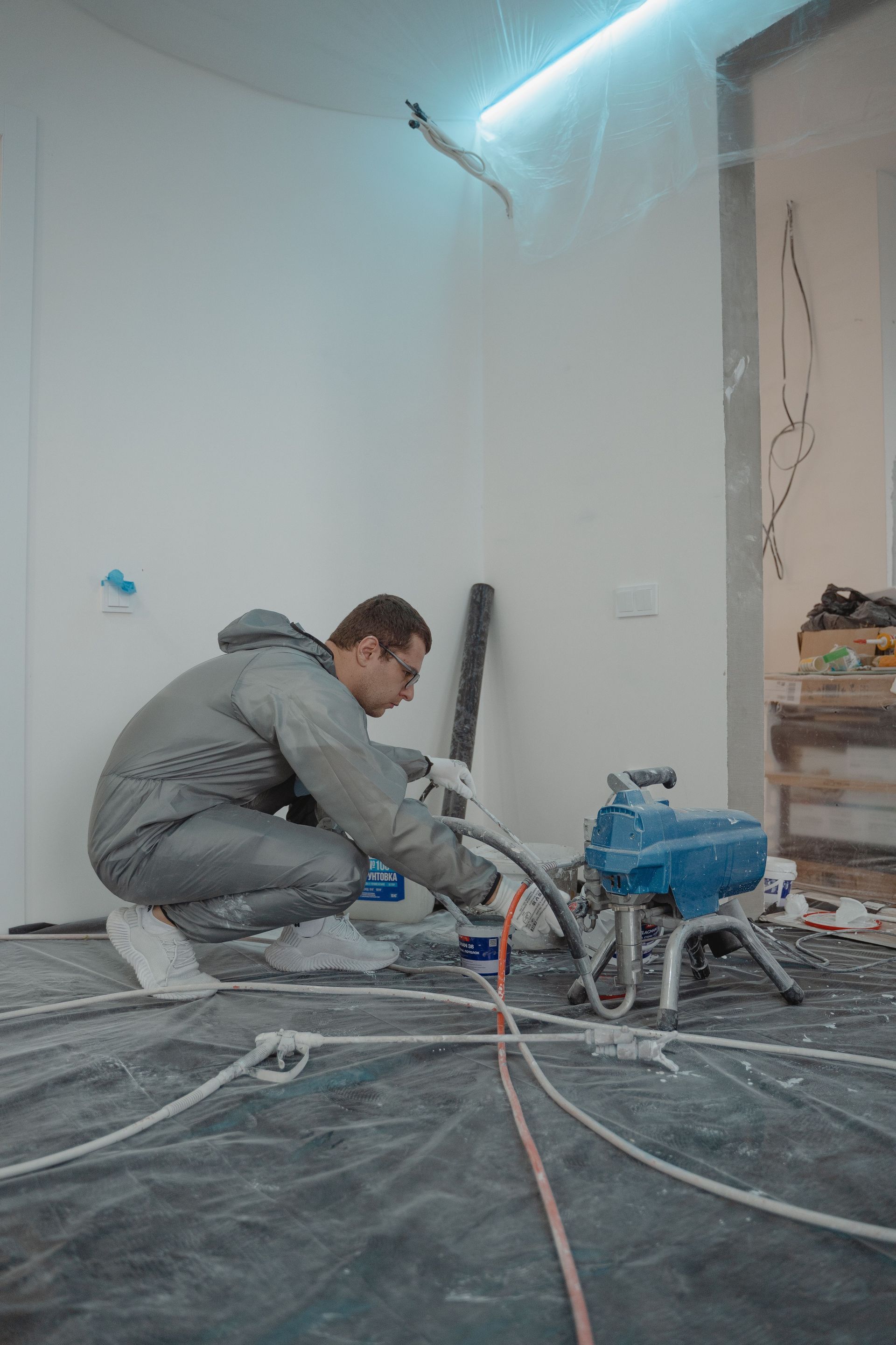 Person crouching in a dusty room using a floor grinder amid renovation debris and cables