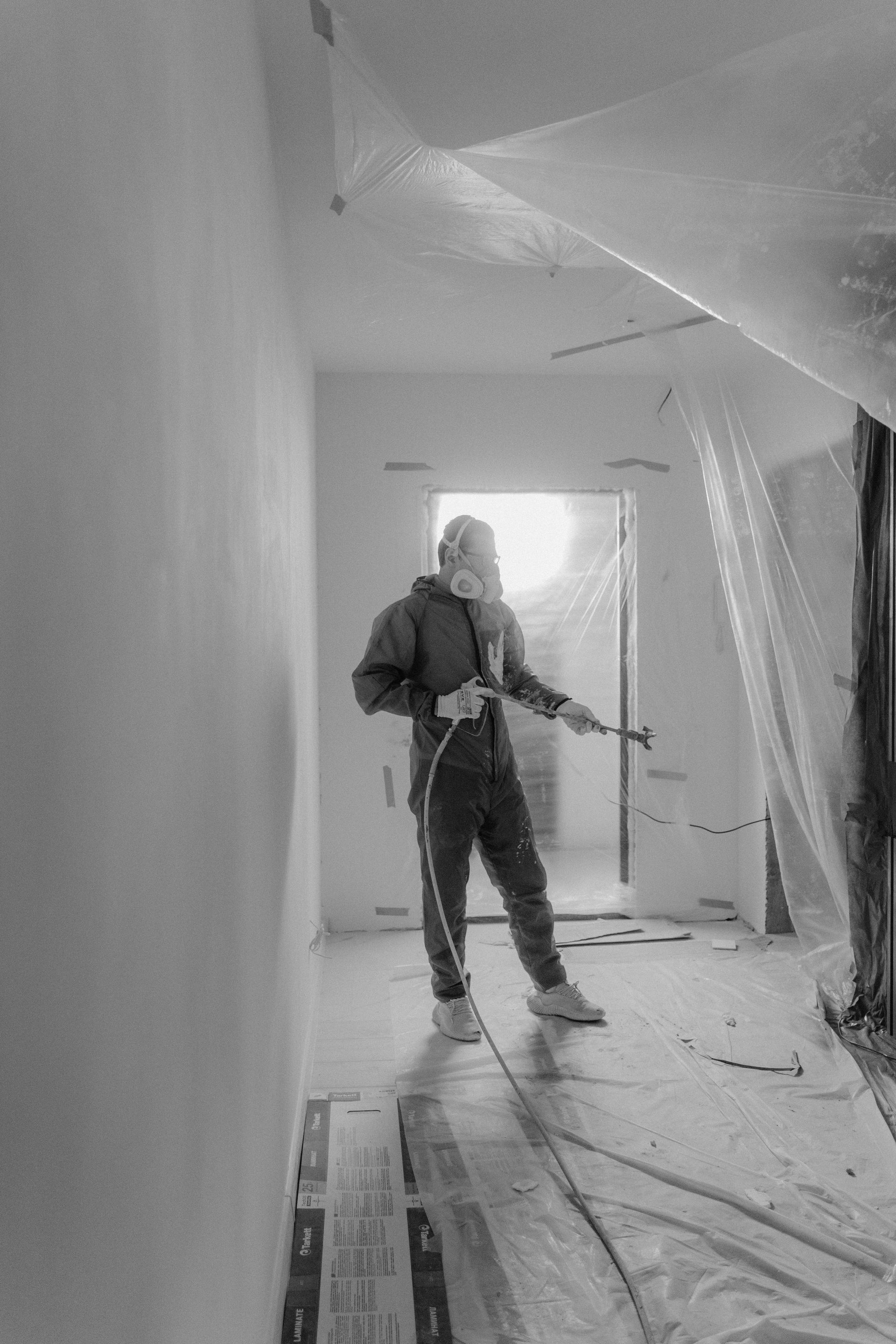 Worker in a narrow hallway using a spray gun during interior painting or renovation.