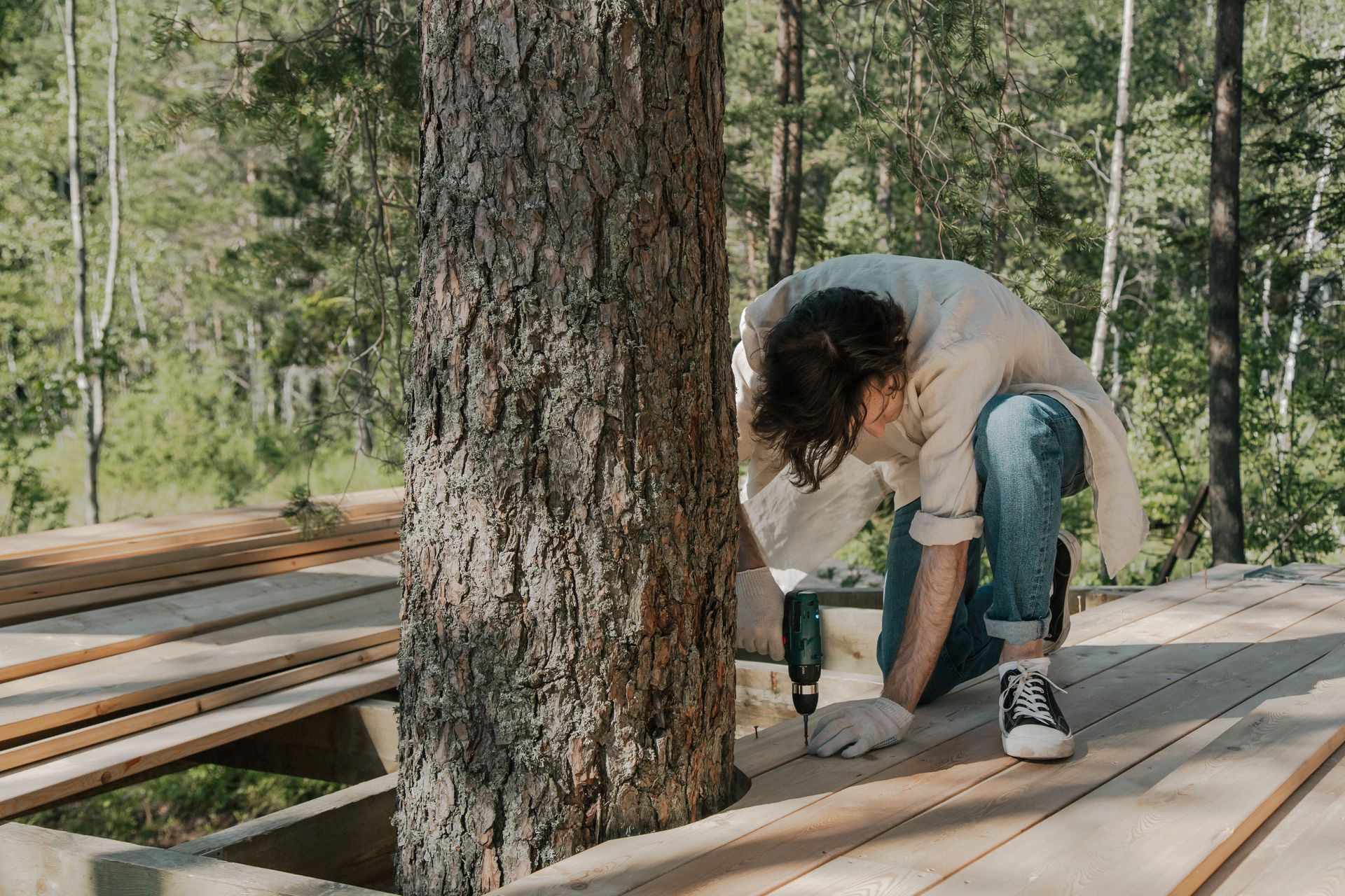 Person drilling a wooden plank beside a tree on a forest deck