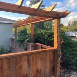 Wooden backyard pergola and fence beside a house, with greenery and a cloudy sky.