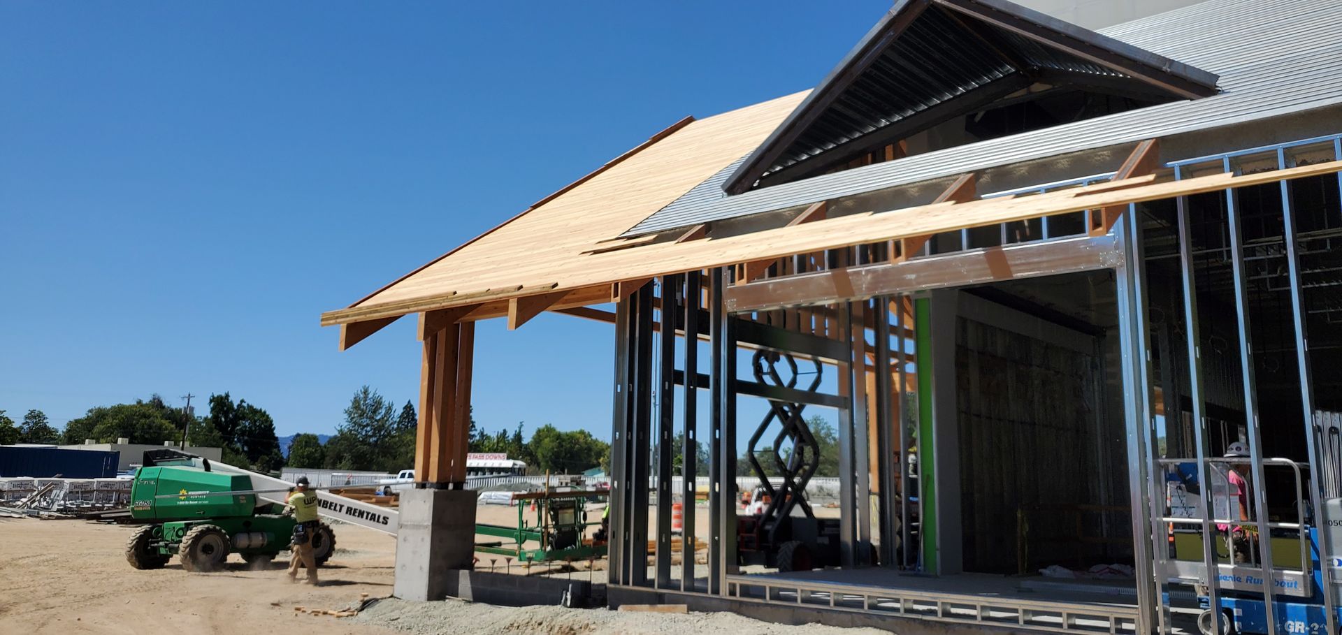 House under construction with exposed framing and roof trusses beside a dirt lot and utility trailer
