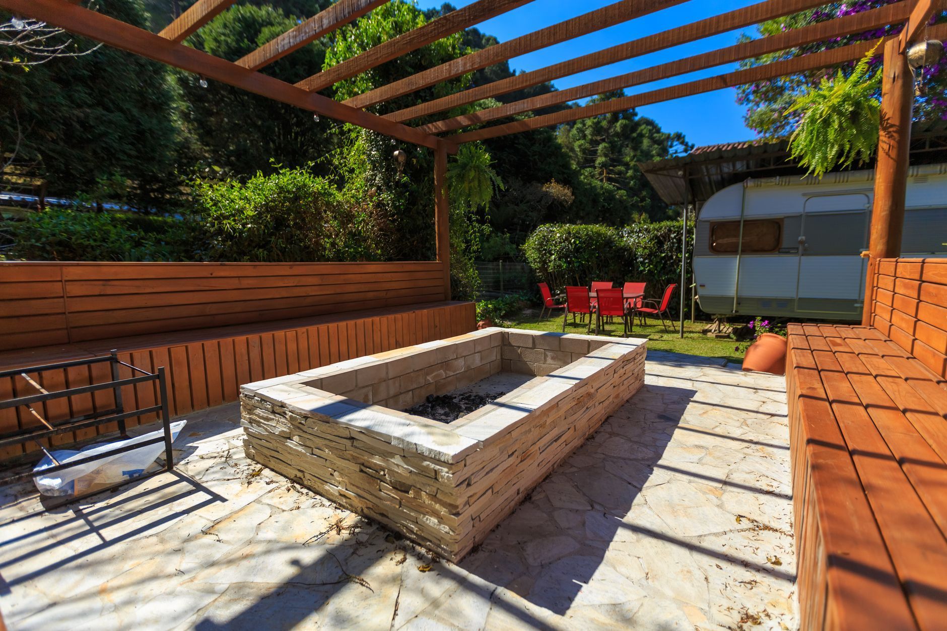 Modern backyard patio with a stone fire pit, wooden benches, pergola, and red chairs in the background