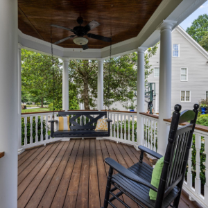 Covered wooden porch with rocking chair, bench swing, white railing, and ceiling fan.