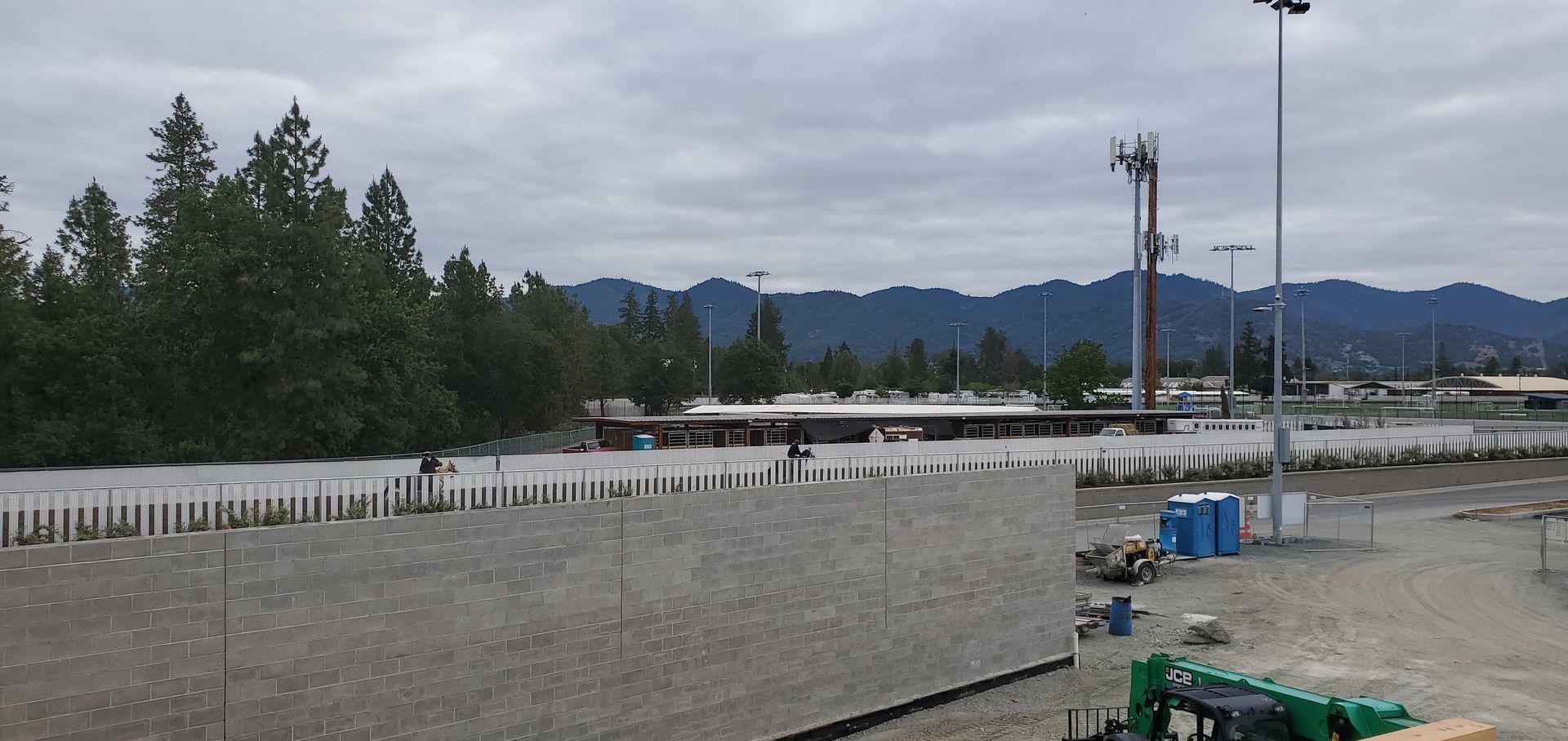 Wide gravel lot beside a white fence, with trees, utility poles, and cloudy mountains in the distance