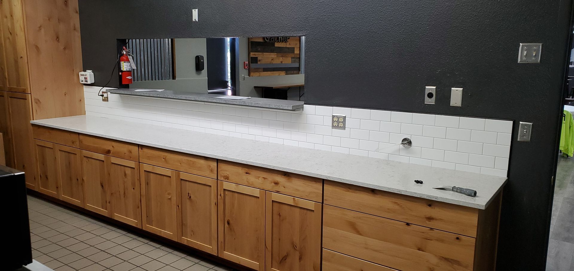 Long countertop with wooden cabinets, white tile backsplash, and dark wall in a commercial kitchen area