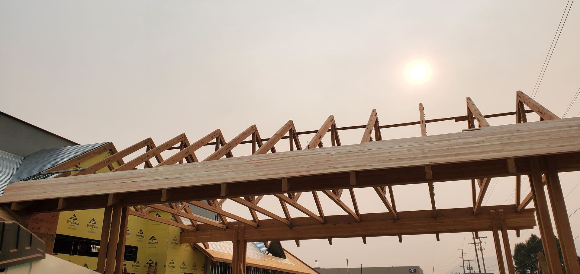 Wooden roof framing under construction at sunset, with a partially covered building and overcast sky.