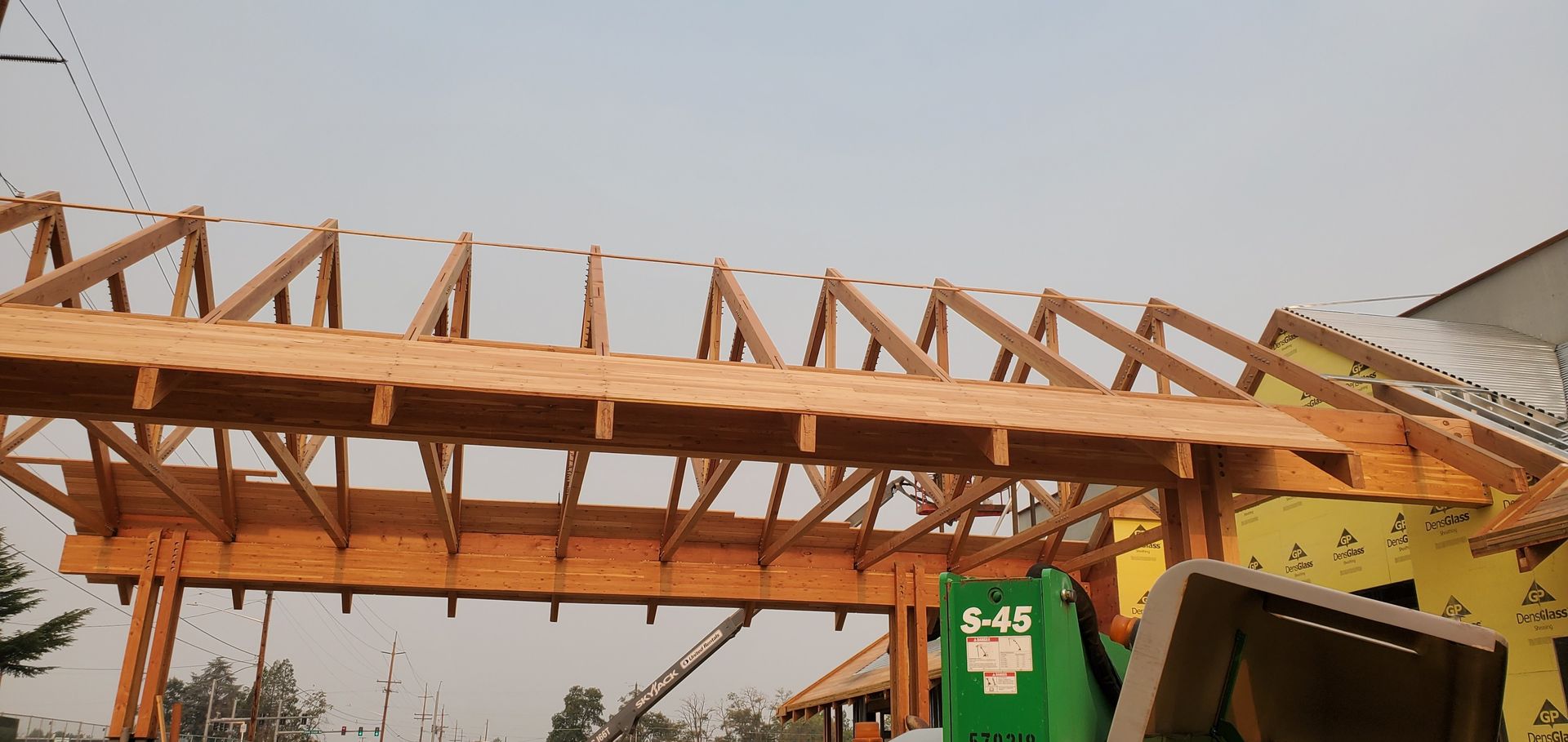Wooden roof trusses under construction beside a green dumpster and a partially visible white building.