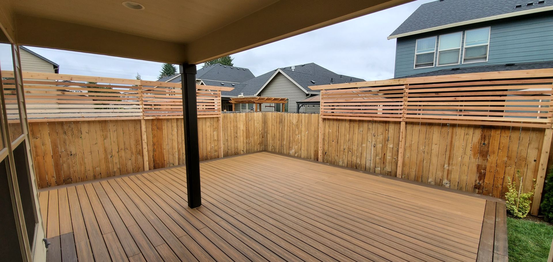 Covered wooden deck with railing, viewed from under a patio roof, overlooking neighboring houses.