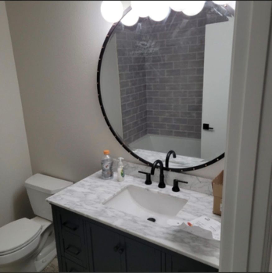 Bathroom vanity with marble sink, round mirror, black faucet, and toilet beside a shower reflected in the mirror