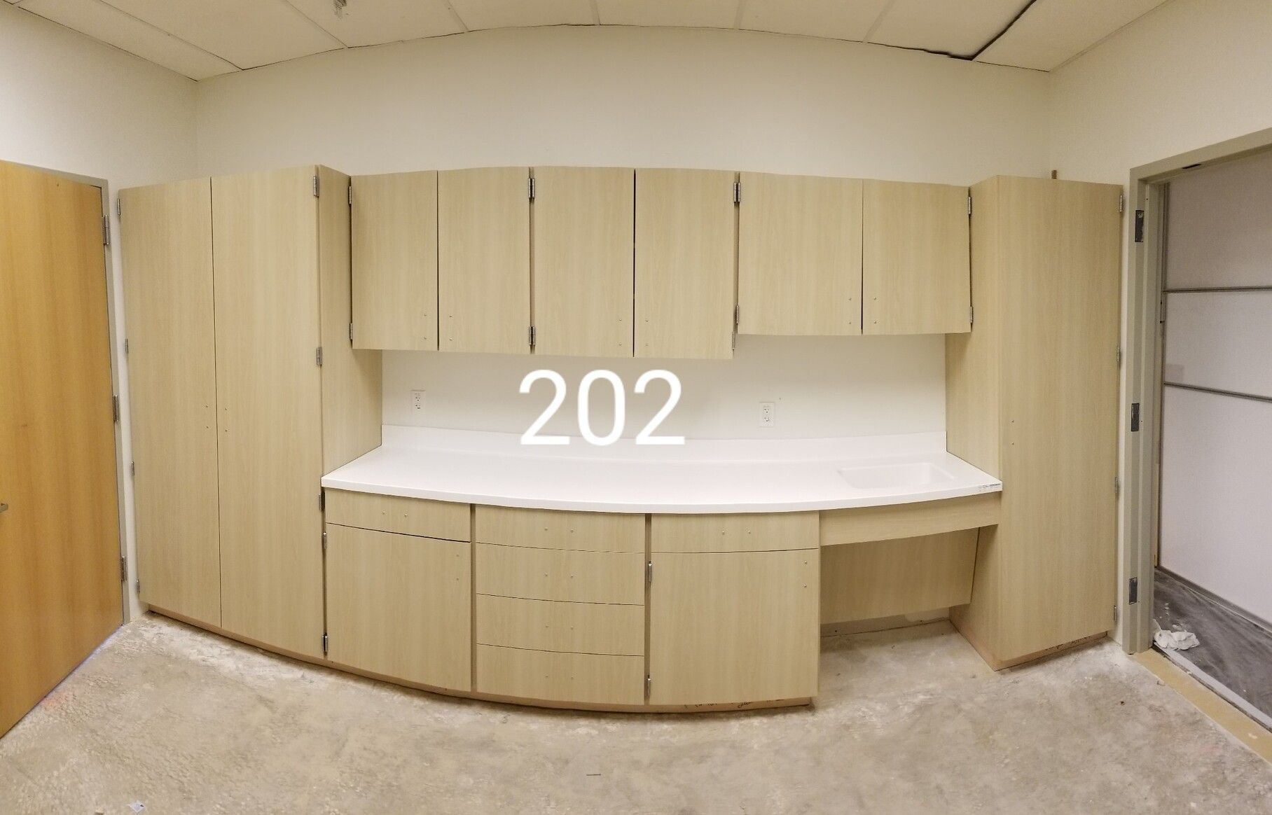 Empty kitchenette with light wood cabinets, white counter, and room number 202 above the sink area.