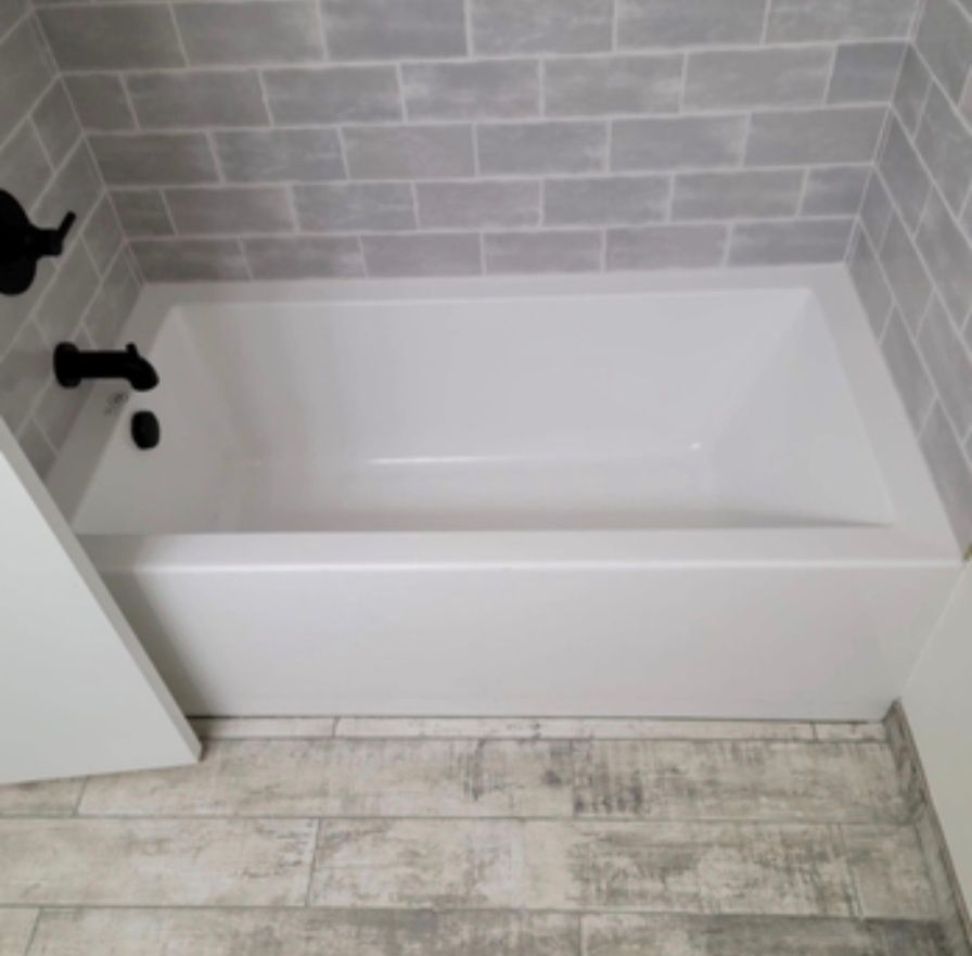 White bathtub with black fixtures in a tiled bathroom, next to gray wood-look flooring