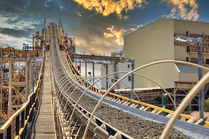 Conveyor belt at a mining facility, transporting material towards a large building under a cloudy sky.