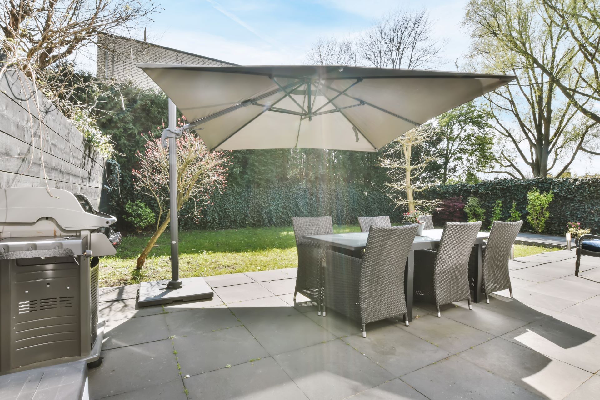 Patio with table, chairs, and umbrella in a backyard with a grill and greenery.