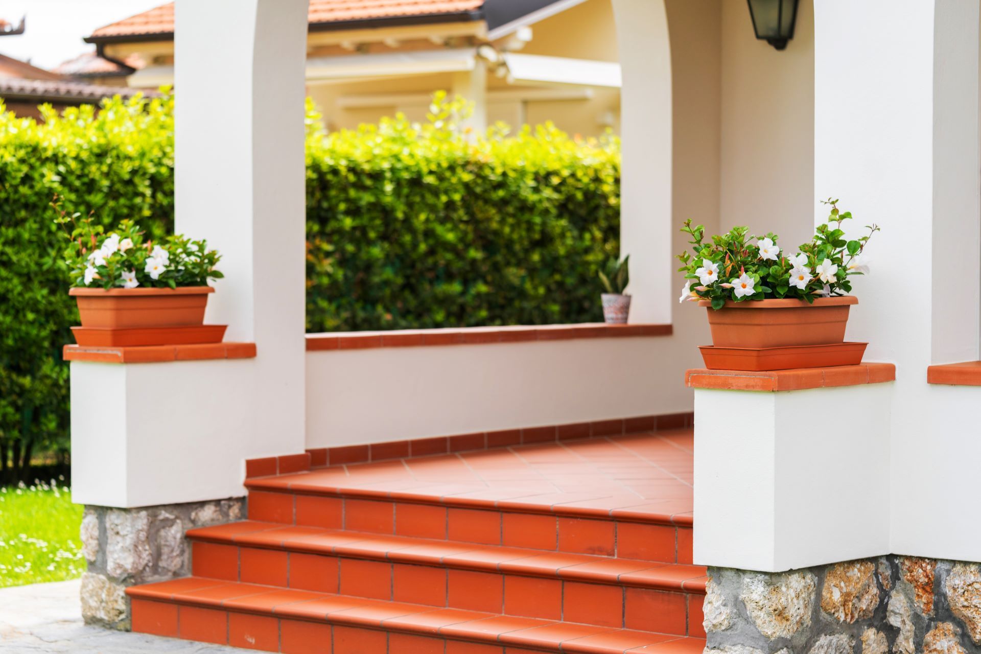 Covered porch with red-tiled steps, white columns, and potted flowers beside a green hedge.