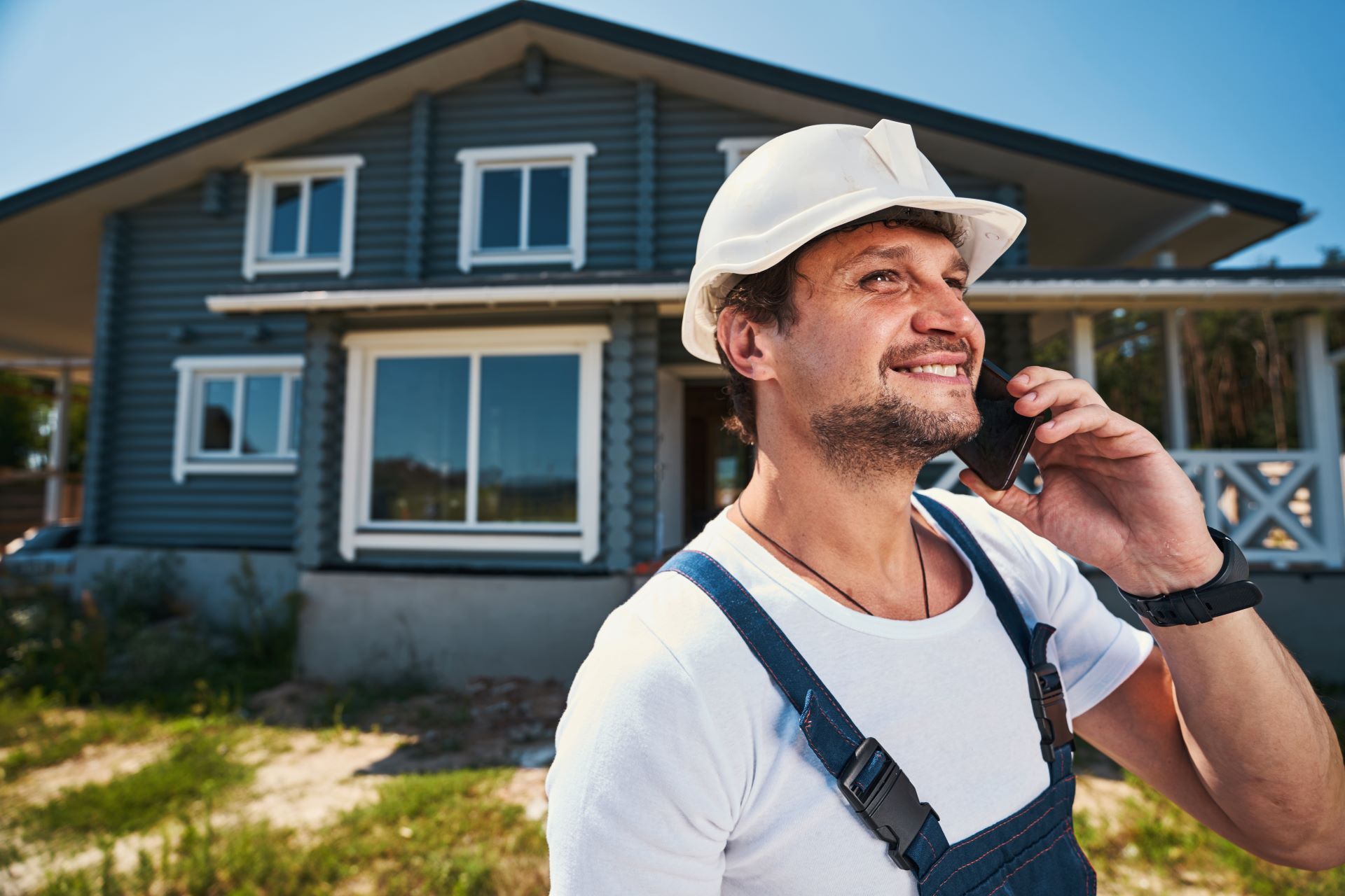 Construction worker in white hard hat smiles while on phone in front of a blue house.