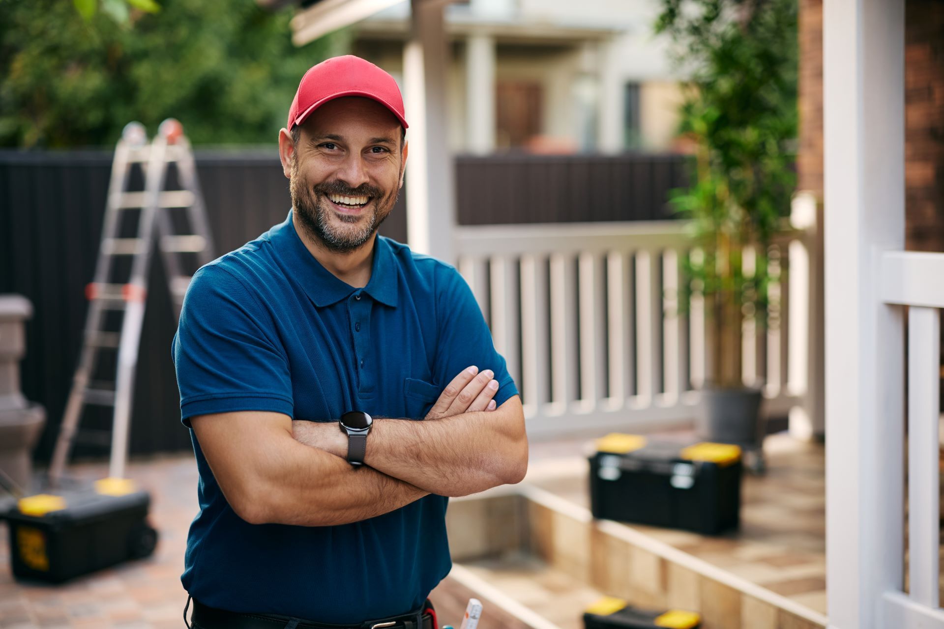 Smiling man in a red cap and blue polo stands with arms crossed in a backyard under renovation
