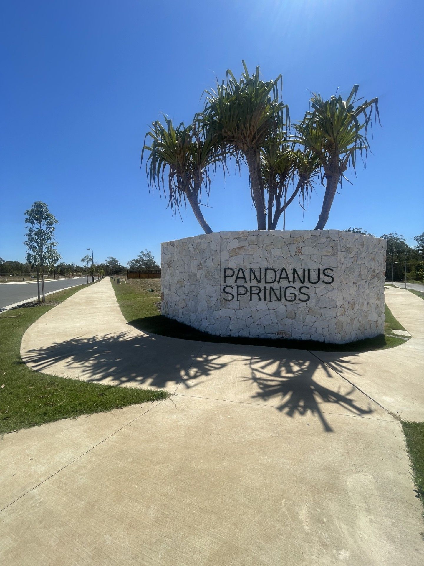 Entrance sign reading “Pandanus Springs” beside a curved walkway and palm trees under a blue sky