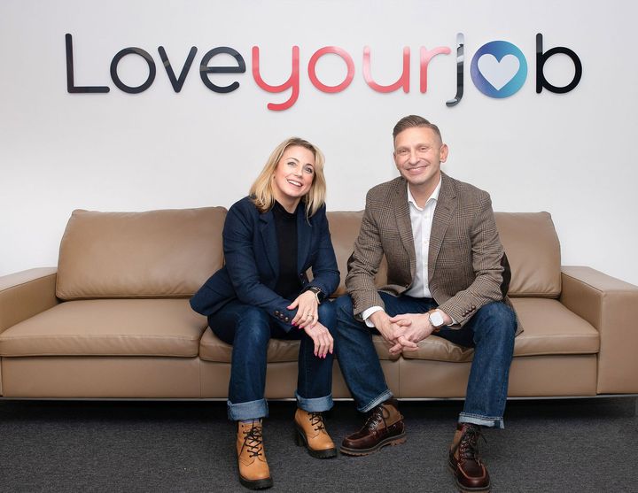 Julie Warner and Haydon Warner sitting on a tan couch beneath a colorful “Loveyourjob” sign.