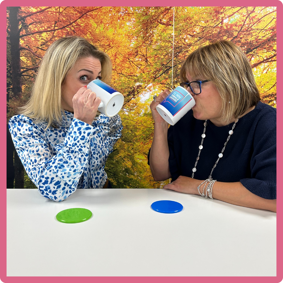 Two women sipping from mugs at a table with autumn foliage background. Green and blue coasters present.