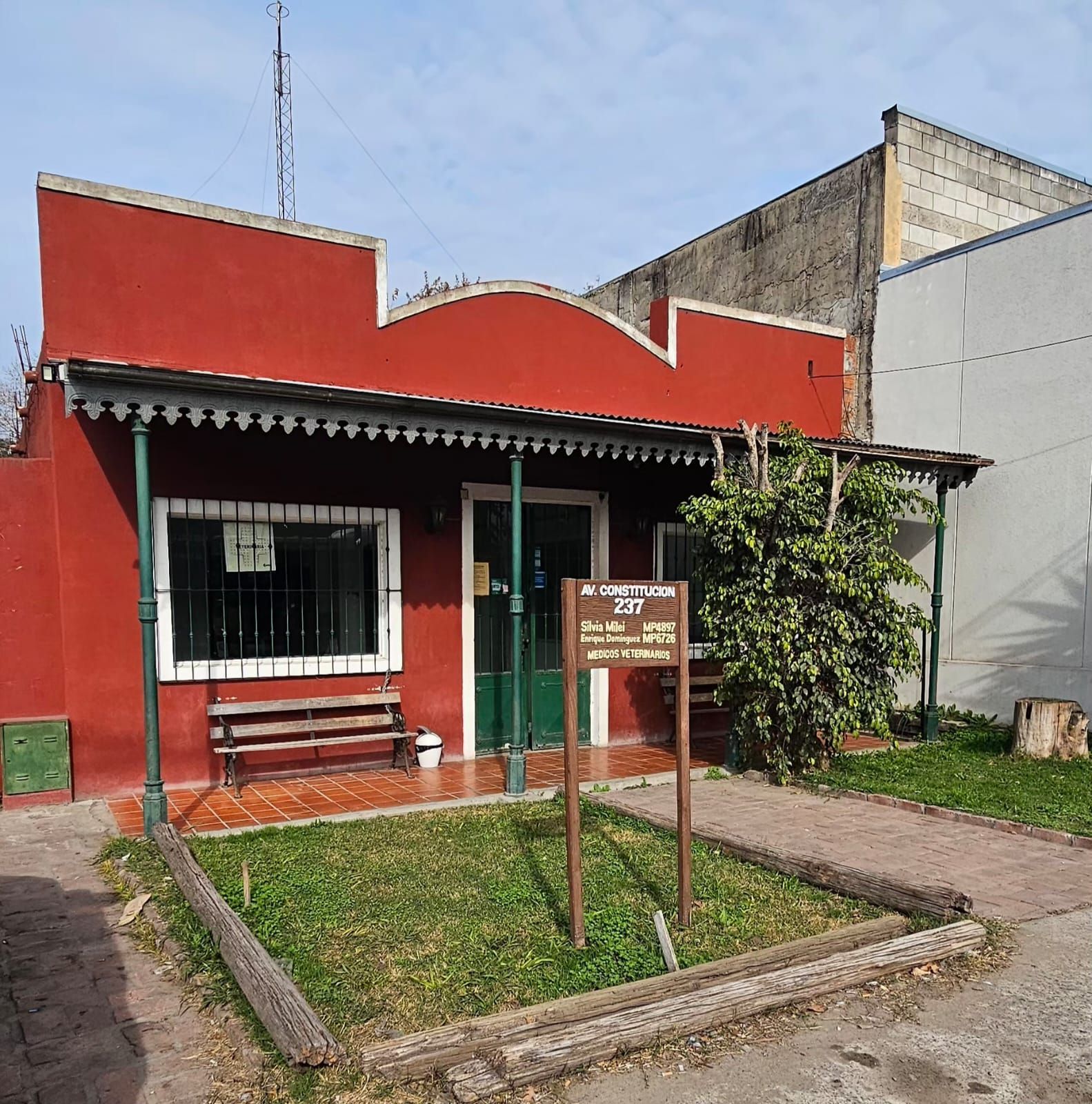 Pequeña casa roja con porche, banco y jardín delantero vallado en una calle soleada.