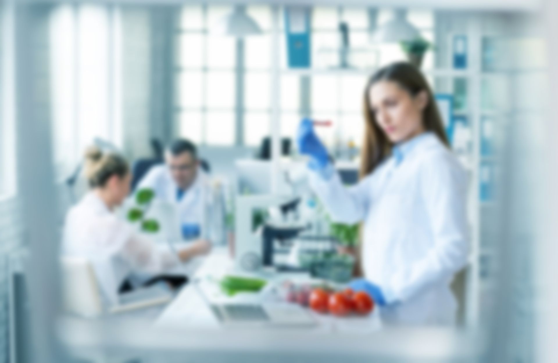 Donna in camice da laboratorio che tiene in mano una provetta con delle verdure davanti, altri scienziati sfocati sullo sfondo.