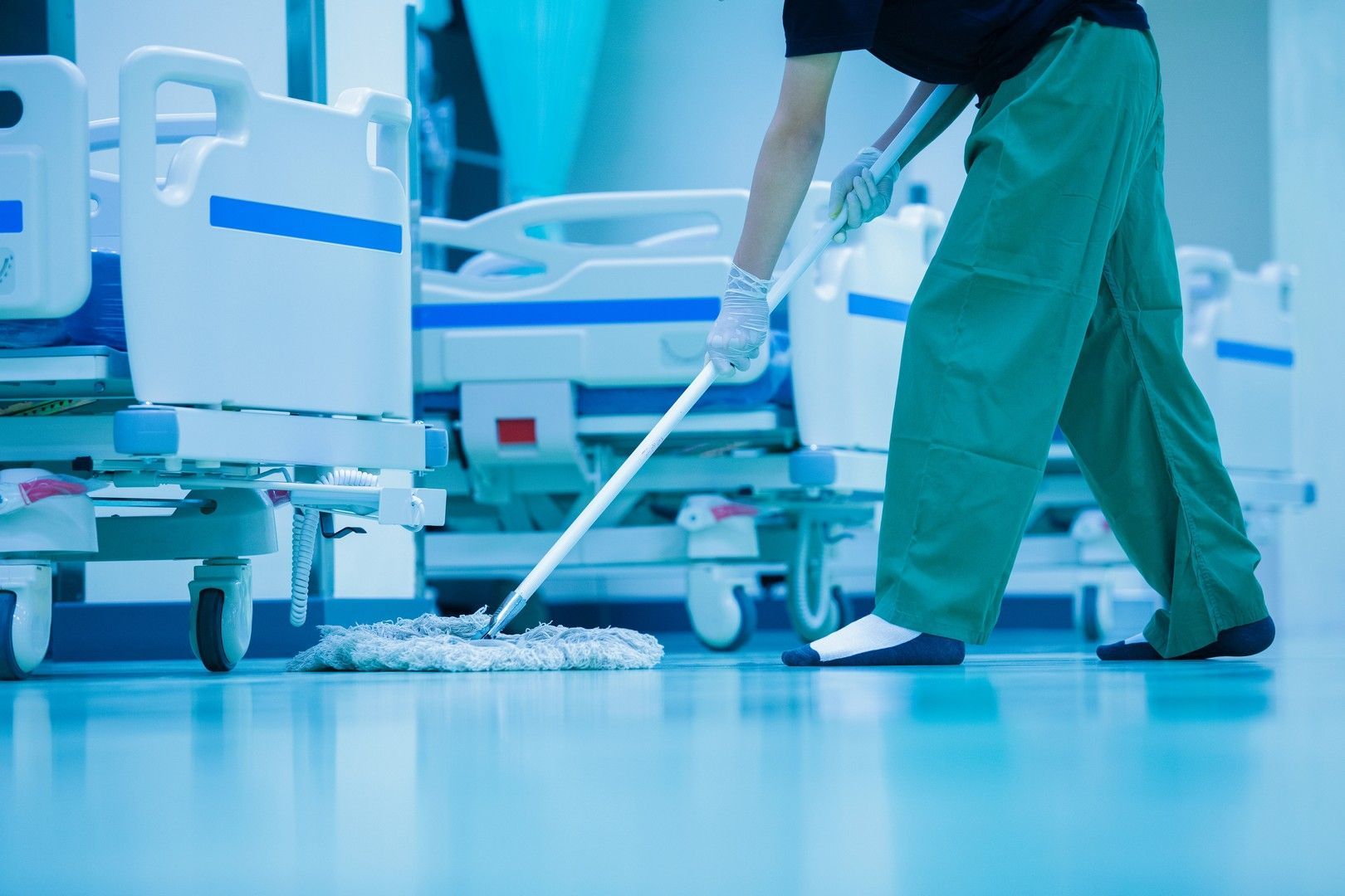 Una persona in camice verde lava i pavimenti in un corridoio dell'ospedale fiancheggiato da letti vuoti.