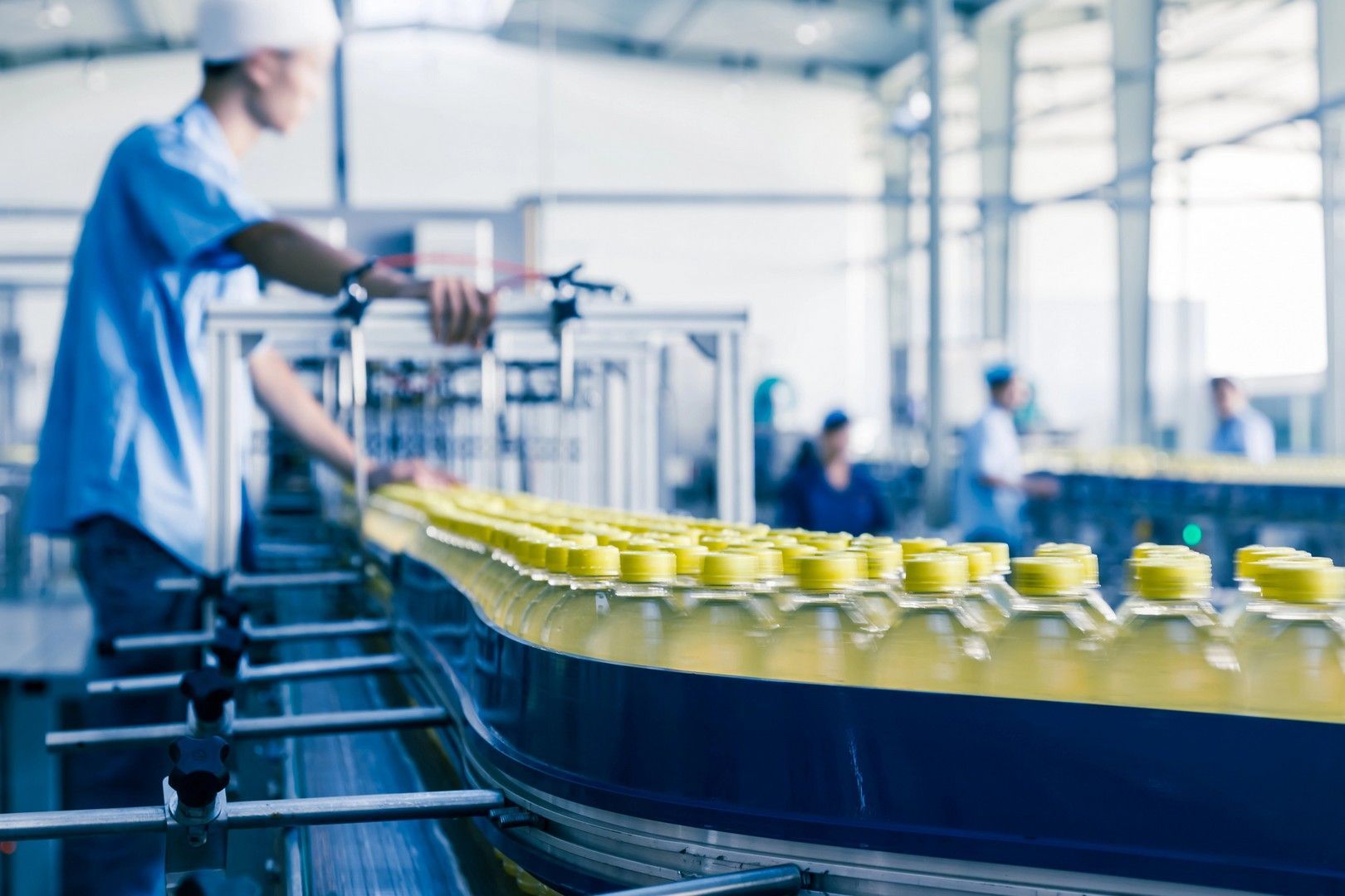 Bottiglie di liquido giallo si muovono lungo un nastro trasportatore in una fabbrica. Un operaio in blu controlla la linea.