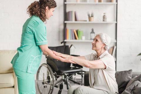 Caregiver in green scrubs holding hands with a seated woman in a bright living room