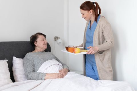 Person in bed smiles as a caregiver offers a tray with fruit in a bright bedroom