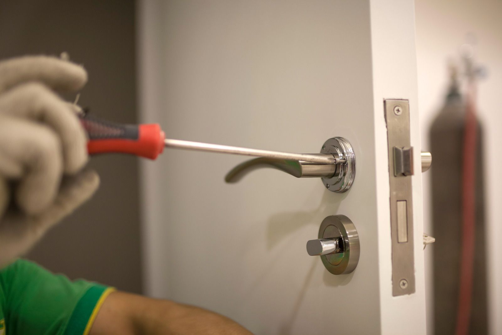 Gloved hand using a red screwdriver to tighten a silver door handle on a white door