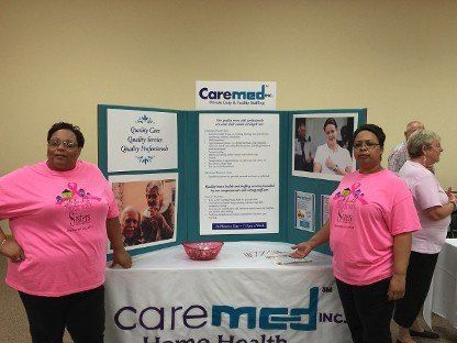 Two women in pink shirts standing at a CareMed information booth with banners and a table.