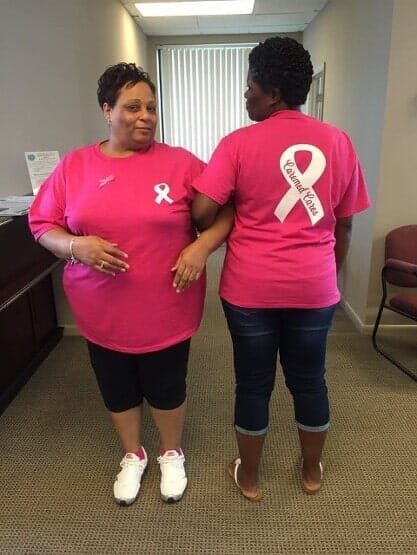 Two women in pink T-shirts with ribbon logos standing indoors, one facing front and one turned away.
