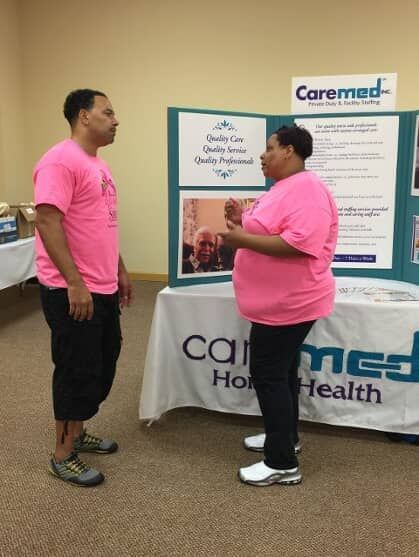 Two people in pink shirts talking at a Caremed Home Health booth with posters and a table display.