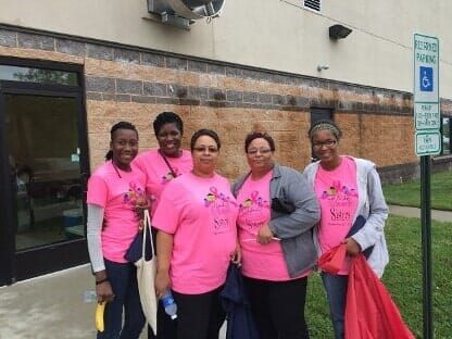 Five people in pink shirts stand outside a building, smiling and posing together.