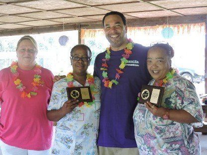 Four people wearing leis hold plaques at a casual indoor celebration.