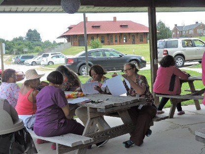 People sitting at a picnic table under a pavilion, with parked cars and a building in the background.