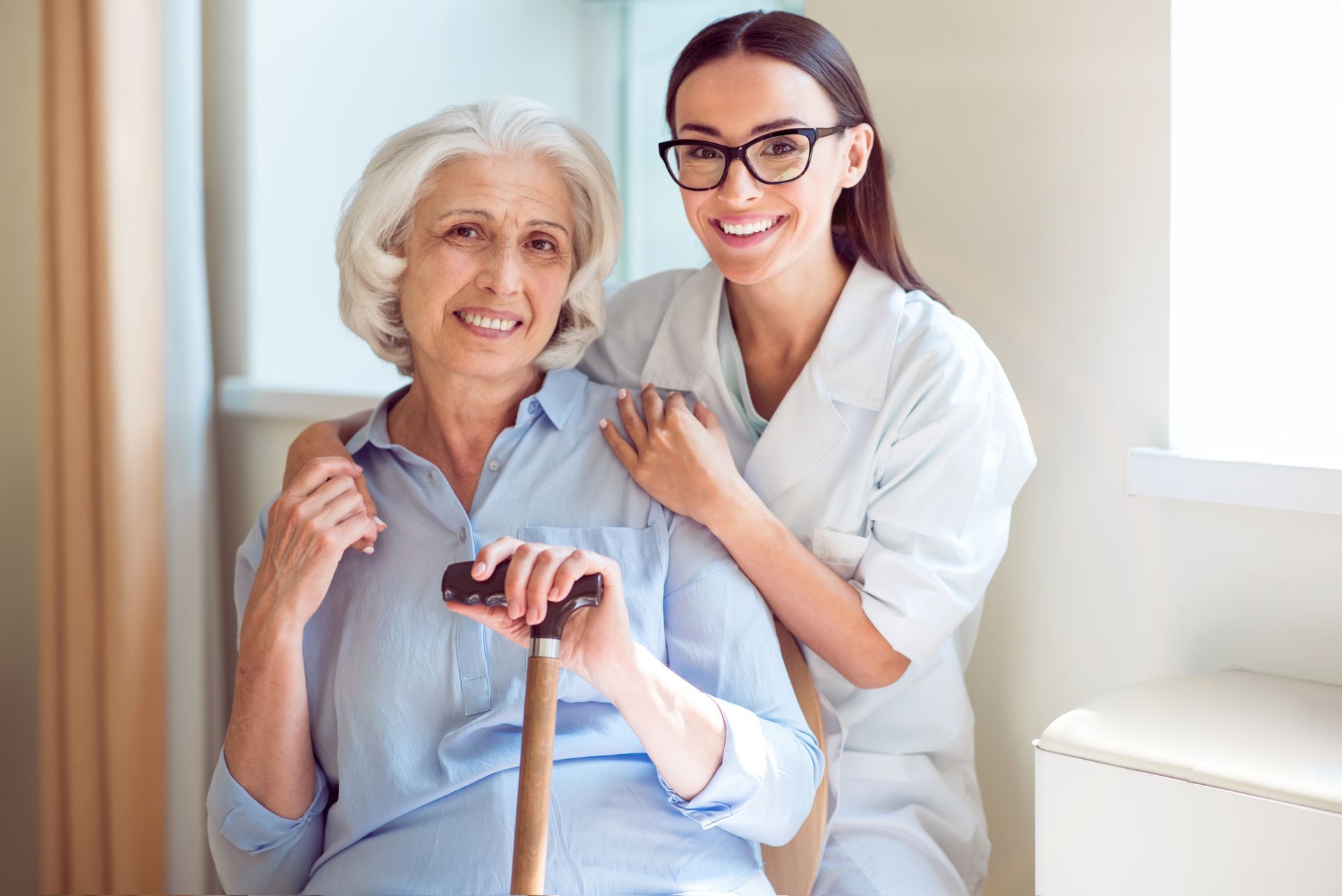Caregiver and elderly woman smiling in a bright room, with the woman holding a cane.