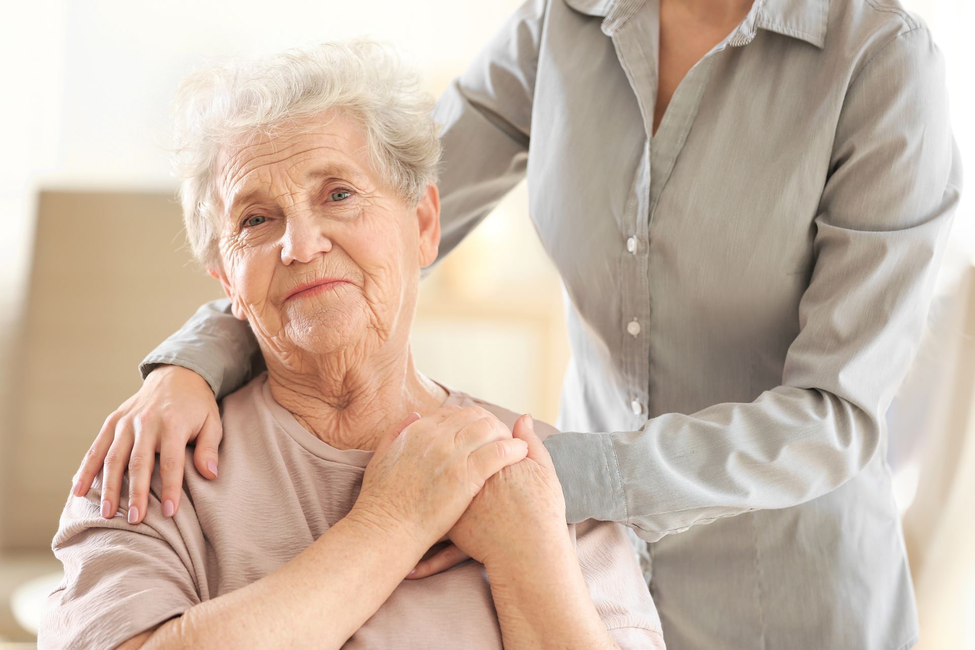 A caregiver gently supports an older woman seated indoors, with a hand on her shoulder.