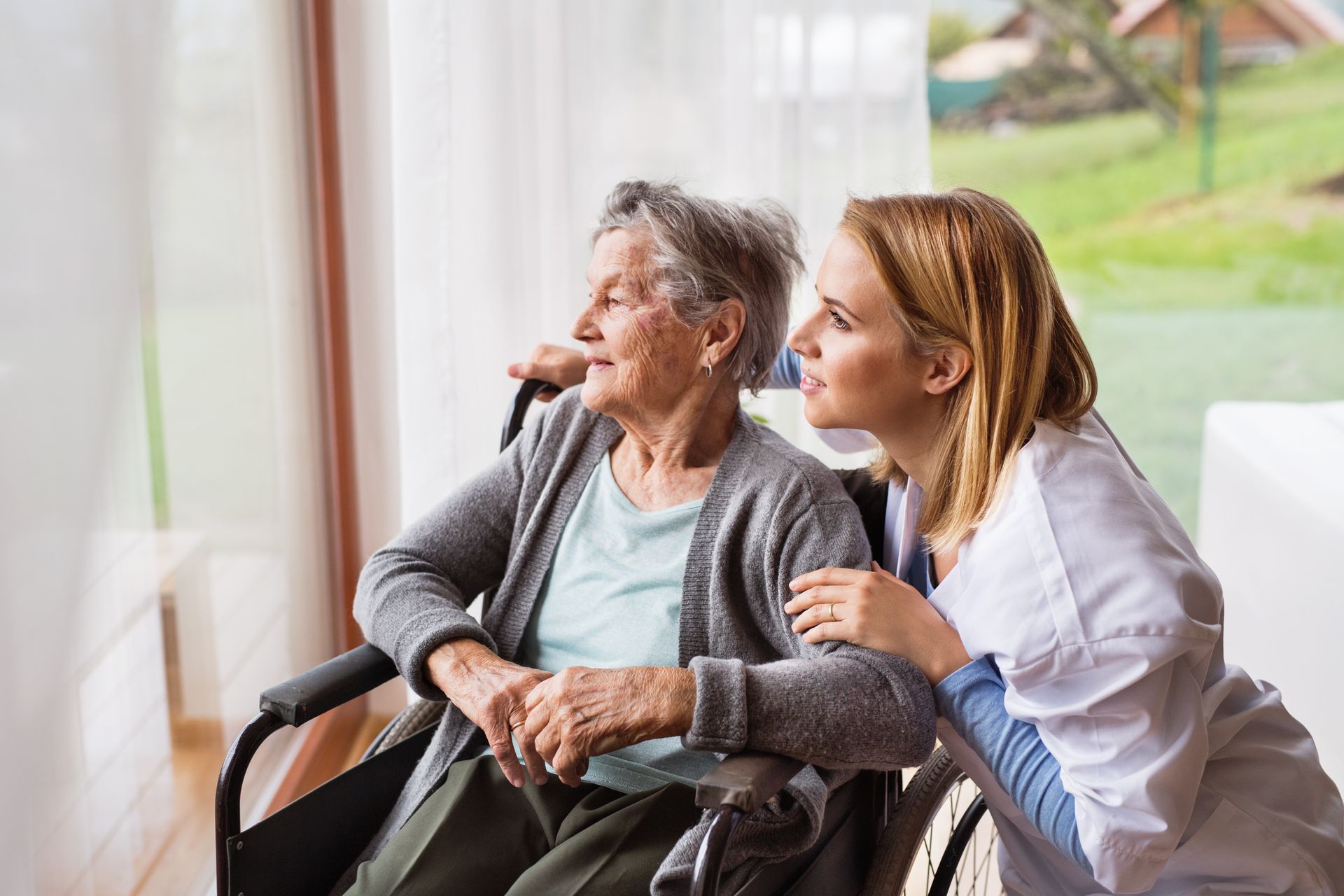 Woman in a wheelchair with a caregiver, sitting by a window and looking outside.