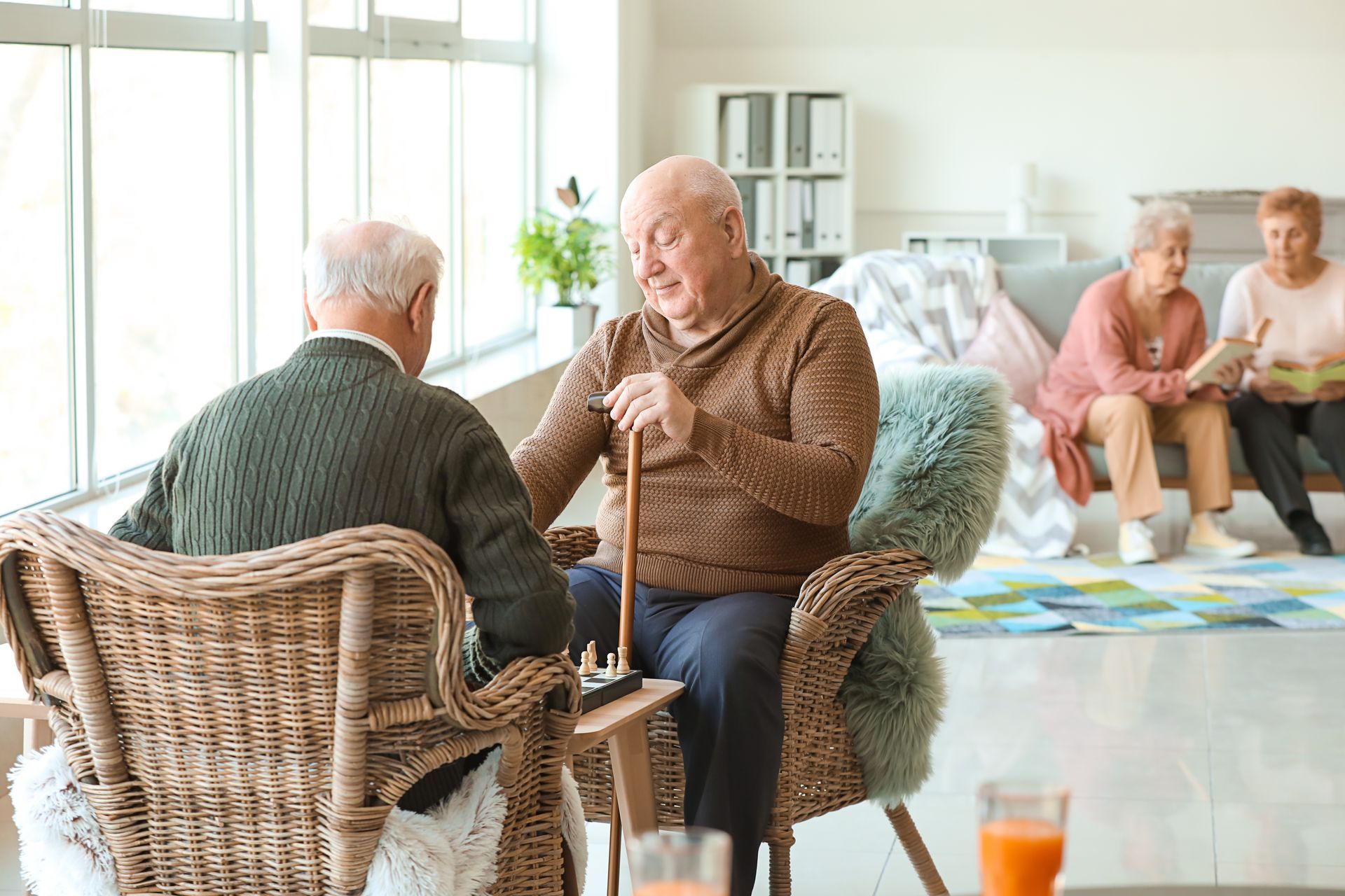 Two seniors sit chatting in wicker chairs in a bright, cozy living room, with others seated in the background.