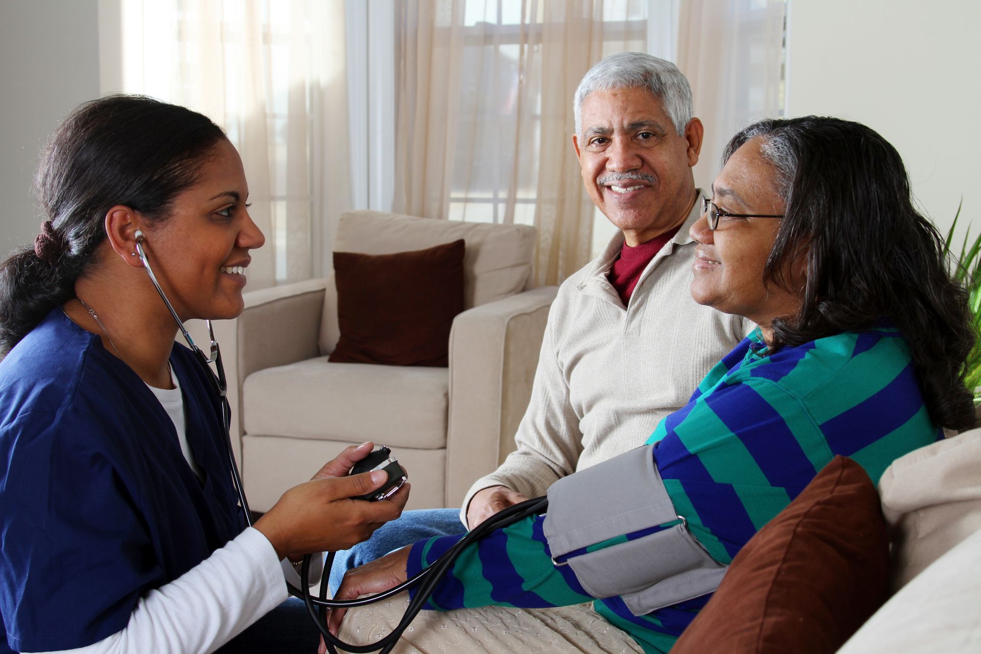 Health professional checking blood pressure for seated patient while another person looks on in a bright living room