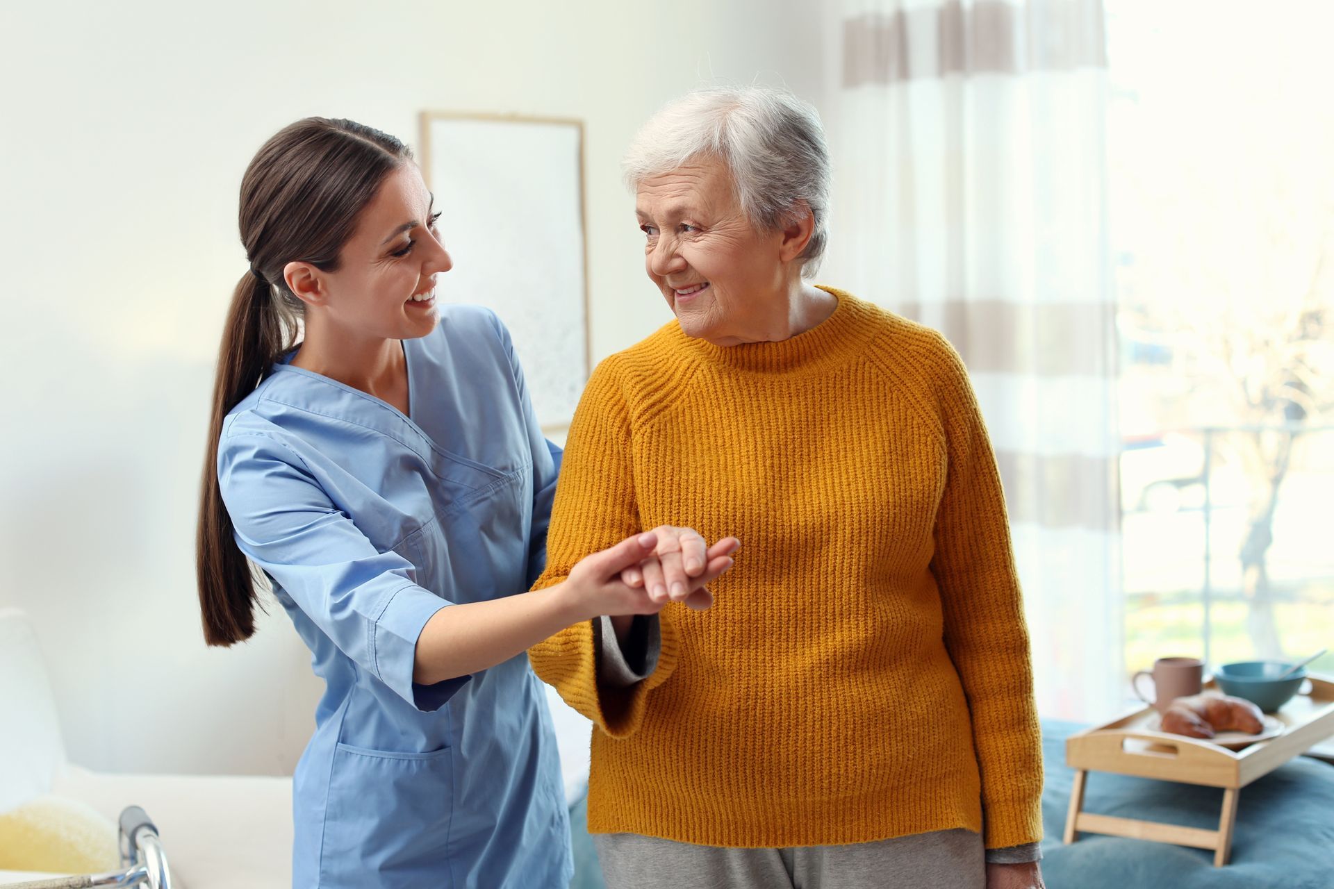 Caregiver in blue scrubs helps a smiling older woman in a yellow sweater stand indoors
